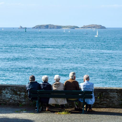 Pięcioro siwych osób w skromnych ubraniach siedzi na ławce naprzeciwko murku przed wybrzeżem. Przed nimi niebieskie wody morza, w oddali jakaś wyspa na skałach